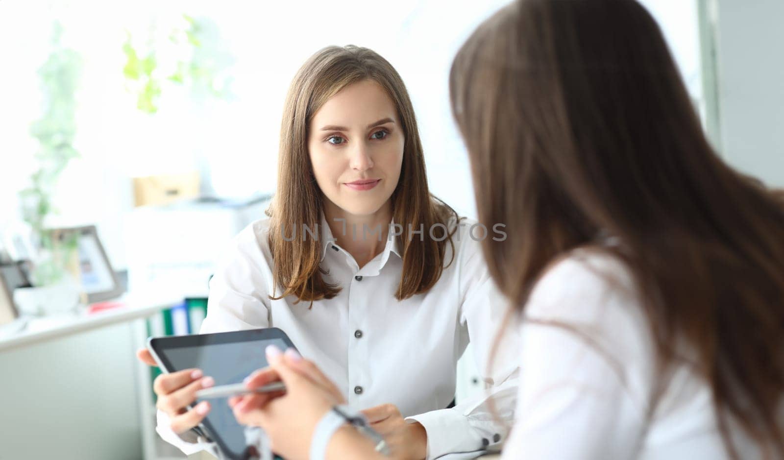 Portrait of joyful businesswoman sitting in office with friendly female manager and holding tablet in tender hands. Copy space on screen. Business meeting concept