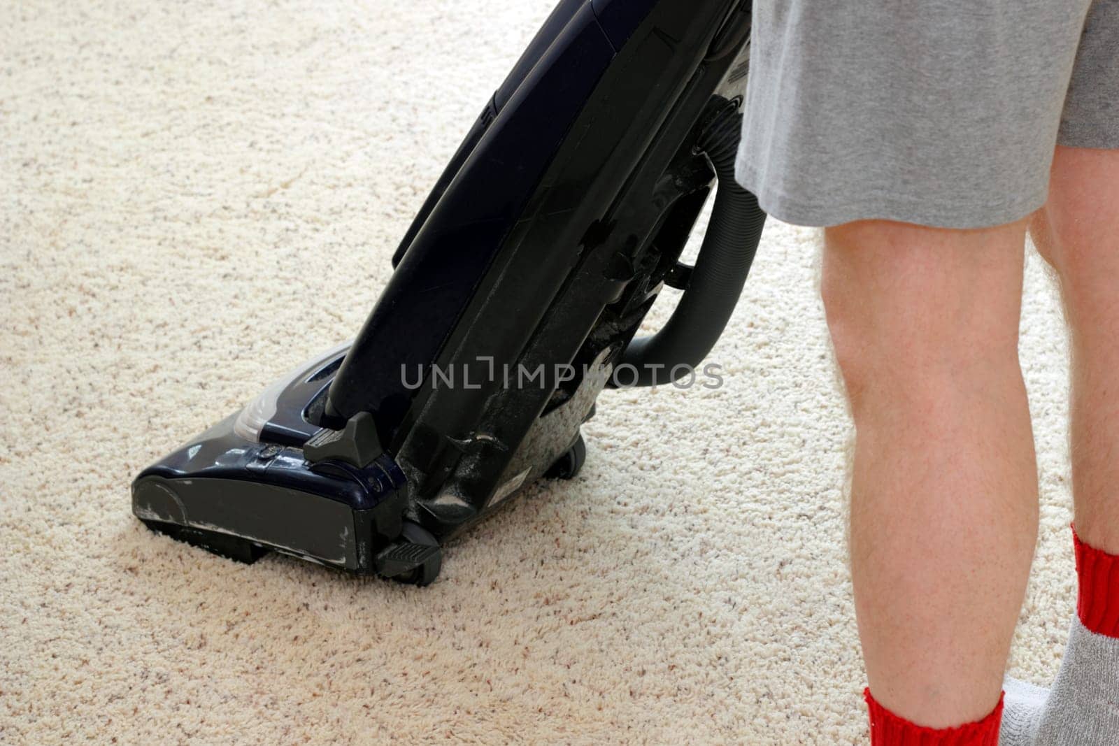 Lower half of a man wearing shorts vacuuming a tan rug with an upright vacuum as part of his housecleaning chores.