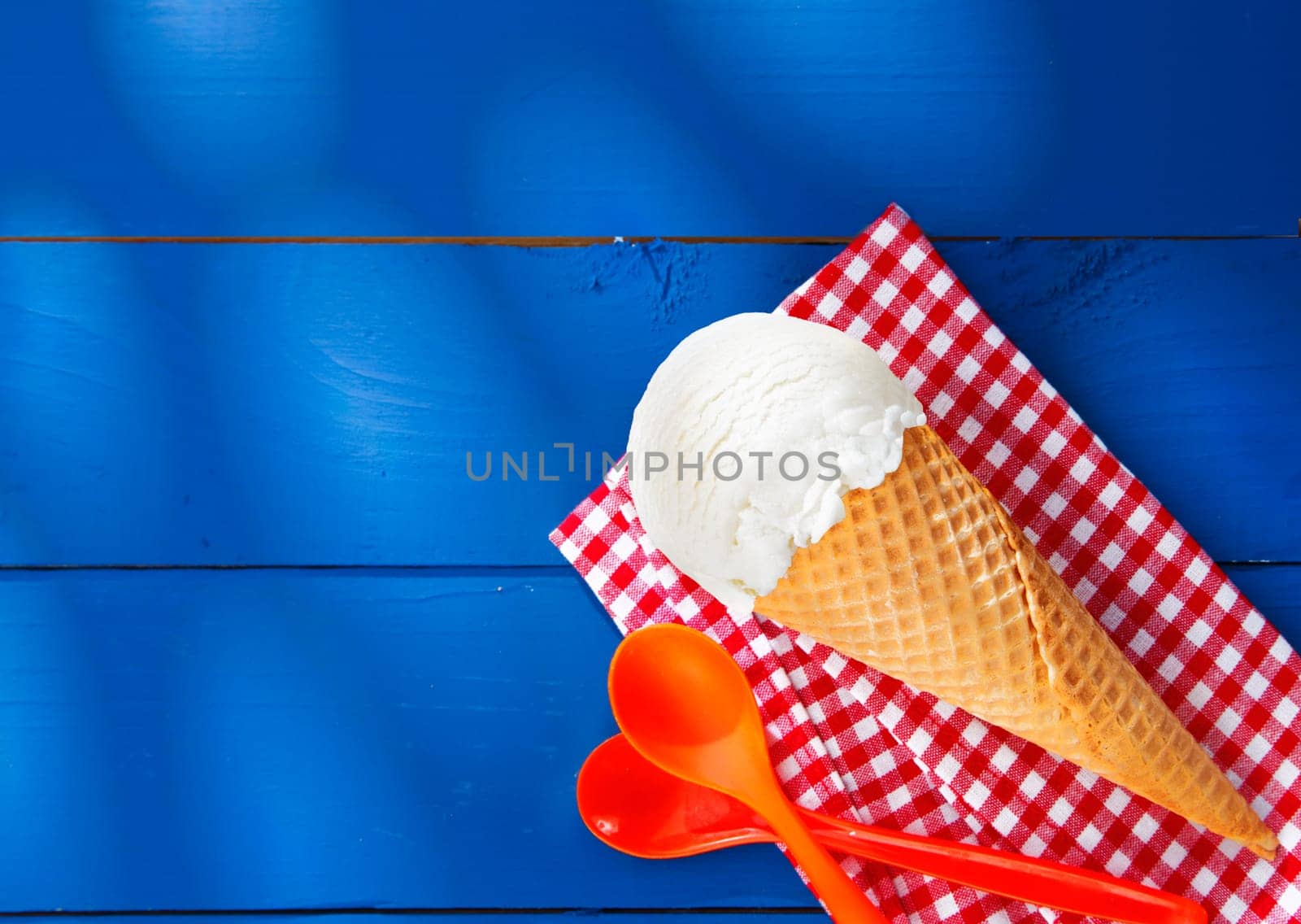 Refreshing creamy vanilla icecream cone served on a red and white checkered napkin lying on rustic blue wooden boards