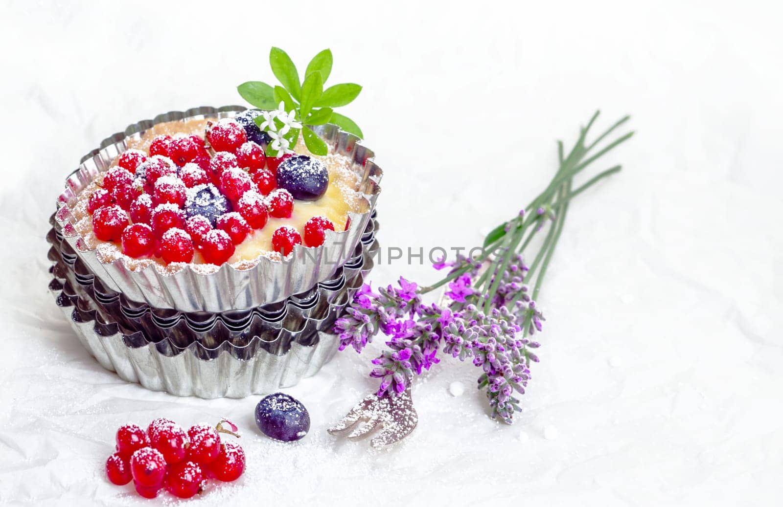 Recurrant tart still life with a freshly baked tart with whole berries balanced on a pile of fluted decorative metal pie pans alongside fresh lavender