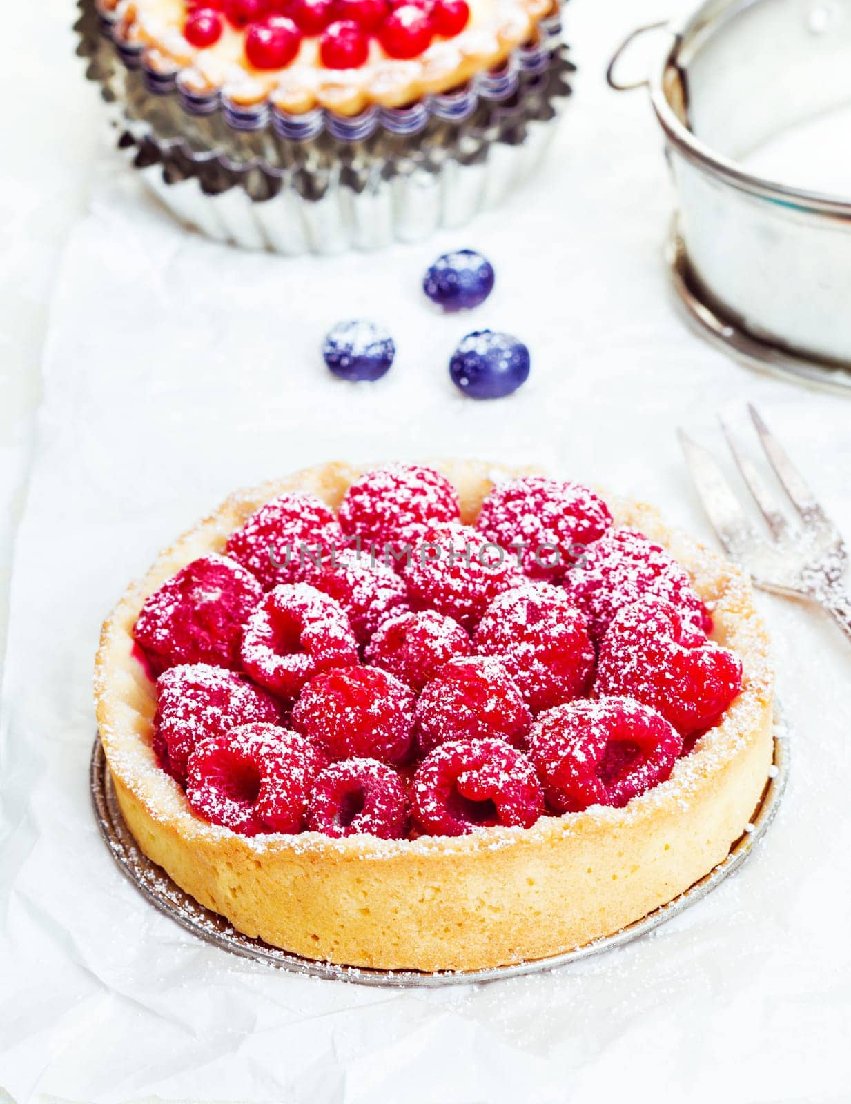Small individual serving of a fresh whole raspberry tart dusted with sugar on a golden crisp crust with a silver cake fork