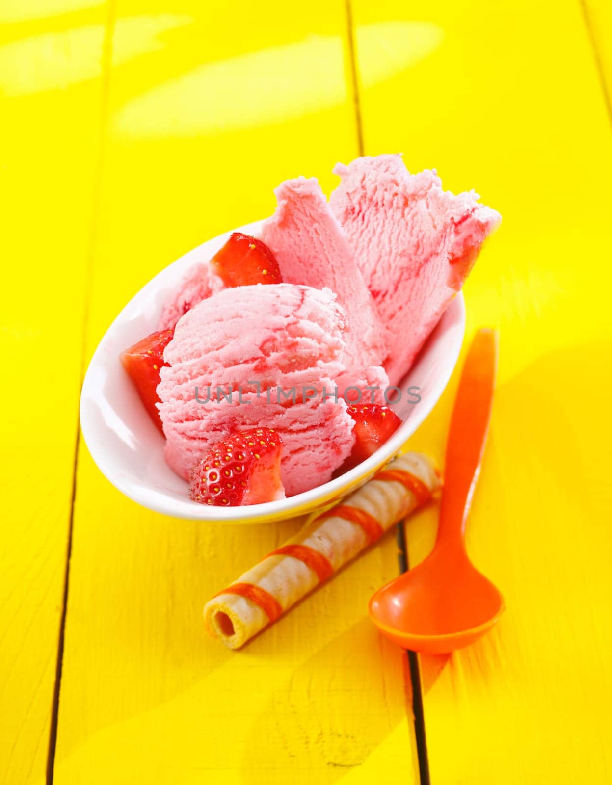 Strawberry icecream with diced ripe red fresh fruit served on a yellow table with a decorative striped wafer biscuit