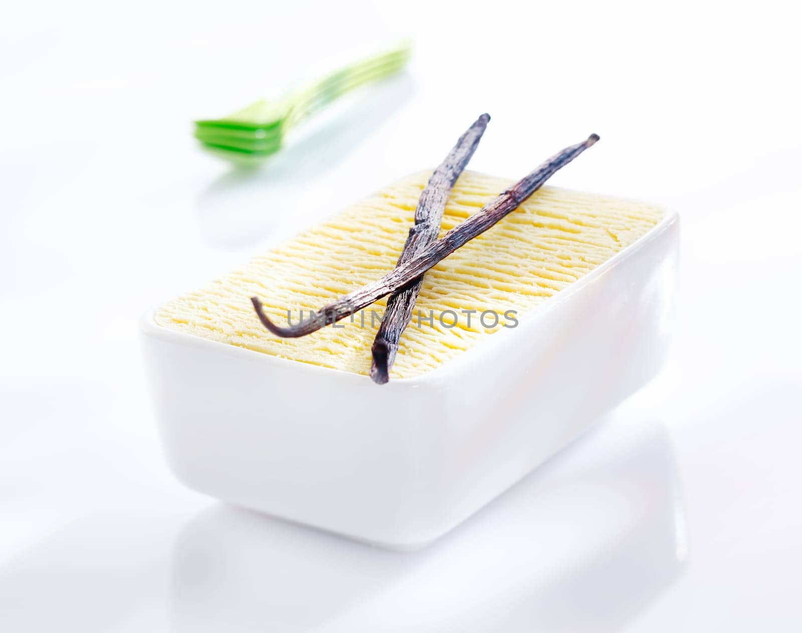 Open tub of creamy vanilla icream with two dried vanilla seedpods used as flavouring in its manufacture on a white background with reflection