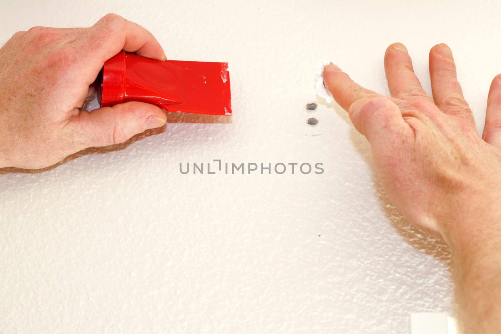 Close up of male hand about to apply white spackling paste to two holes in the wall while holding on other hand a red tool to smooth spackling.