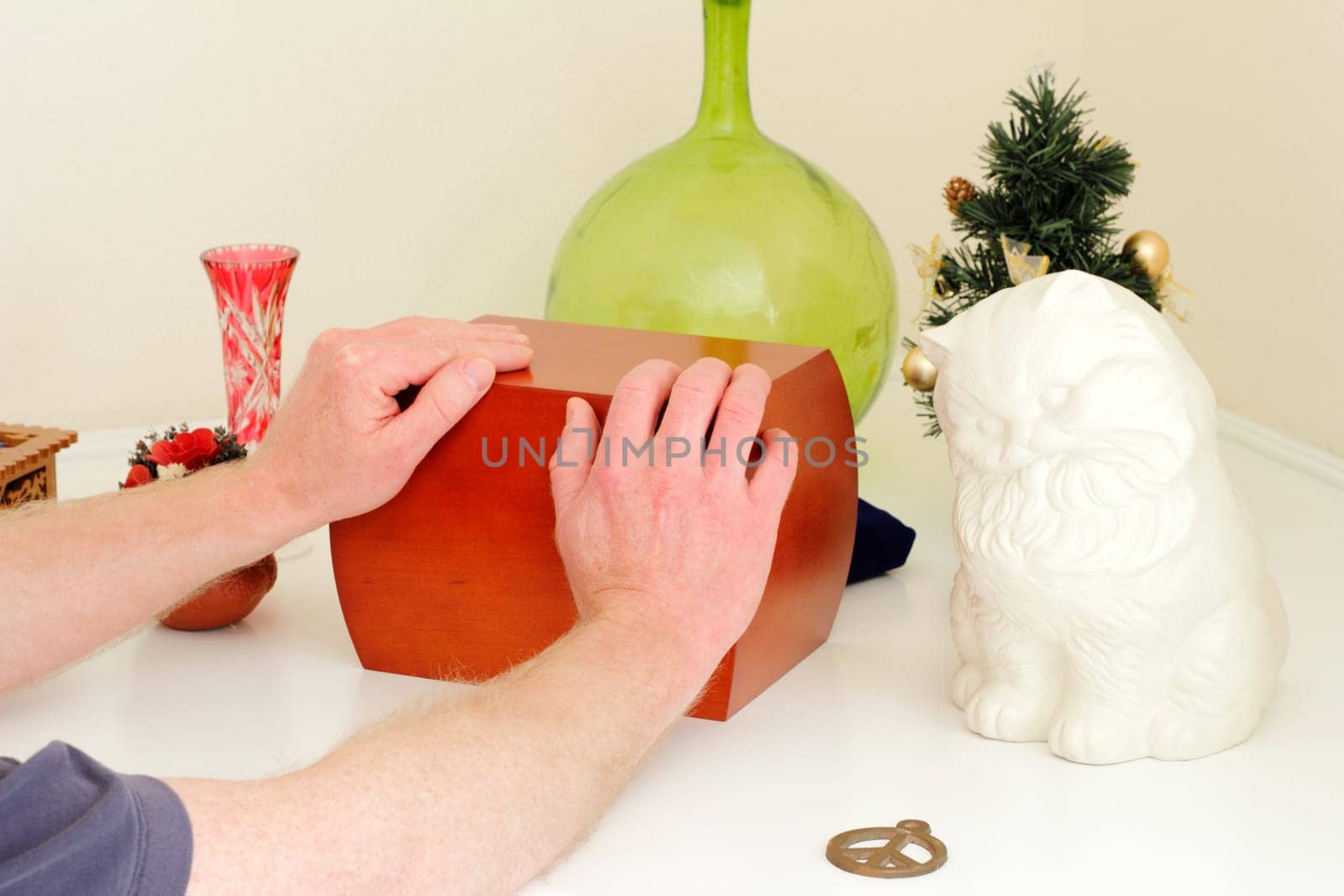 Male caucasian hands holding a wood urn that is on a white mantle surrounded by the departed loved ones mementoes.