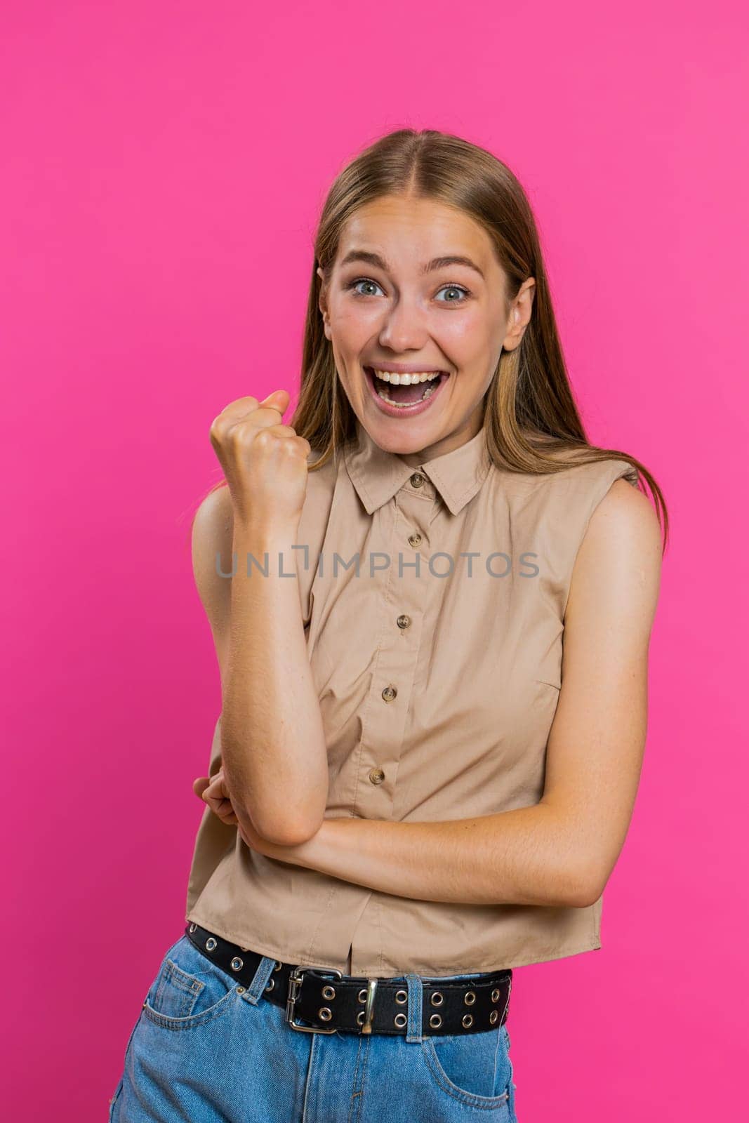 Happy joyful woman shouting, raising fists in gesture I did it, celebrating success, winning, birthday, lottery jackpot goal achievemen, good news. Young girl on pink studio wall background. Vertical