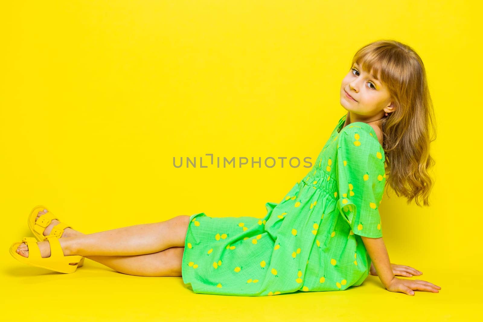Girl kid in green dress lying on floor, relaxing, chill, enjoying, looking at camera with smile. Preteen Caucasian child having fun, posing isolated on bright shine vibrant yellow background
