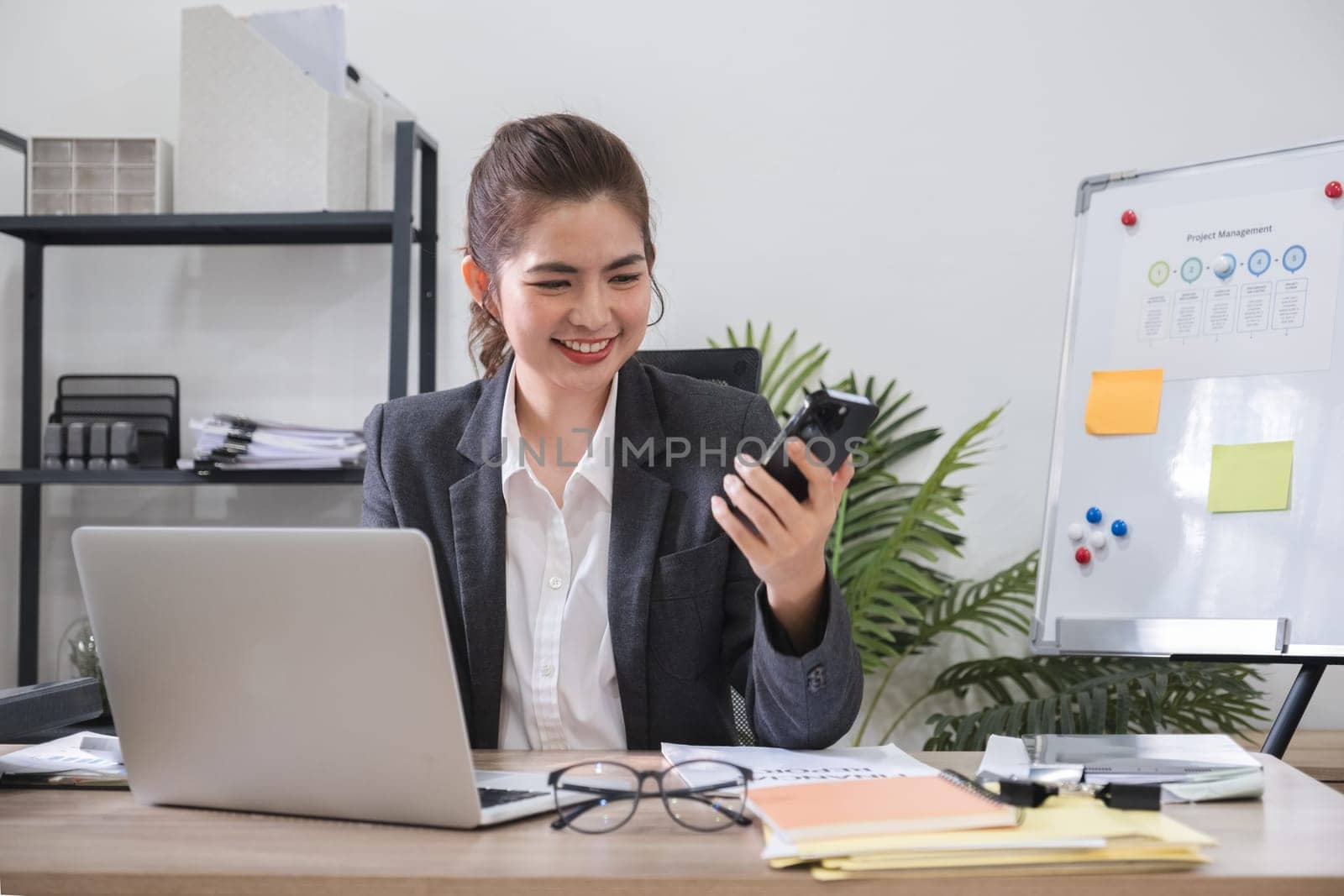 Asian female office worker with bright smile video chats on smartphone with business team, planning work and corporate marketing..