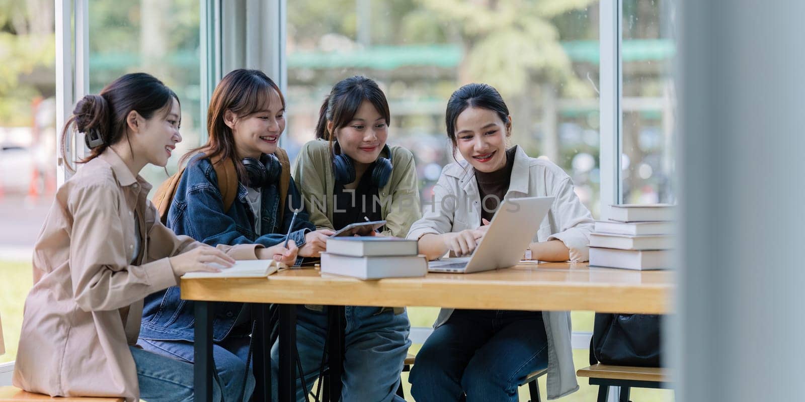 Group of young Asian college students sitting on a bench in a campus relaxation area, talking, sharing ideas, doing homework or tutoring for the exam together.
