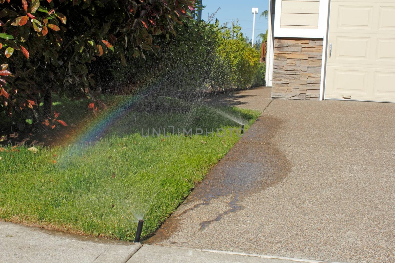 On the front left side of a home a lawn sprinkler is spraying water and creating a rainbow over the grass on a sunny blue sky day in spring.