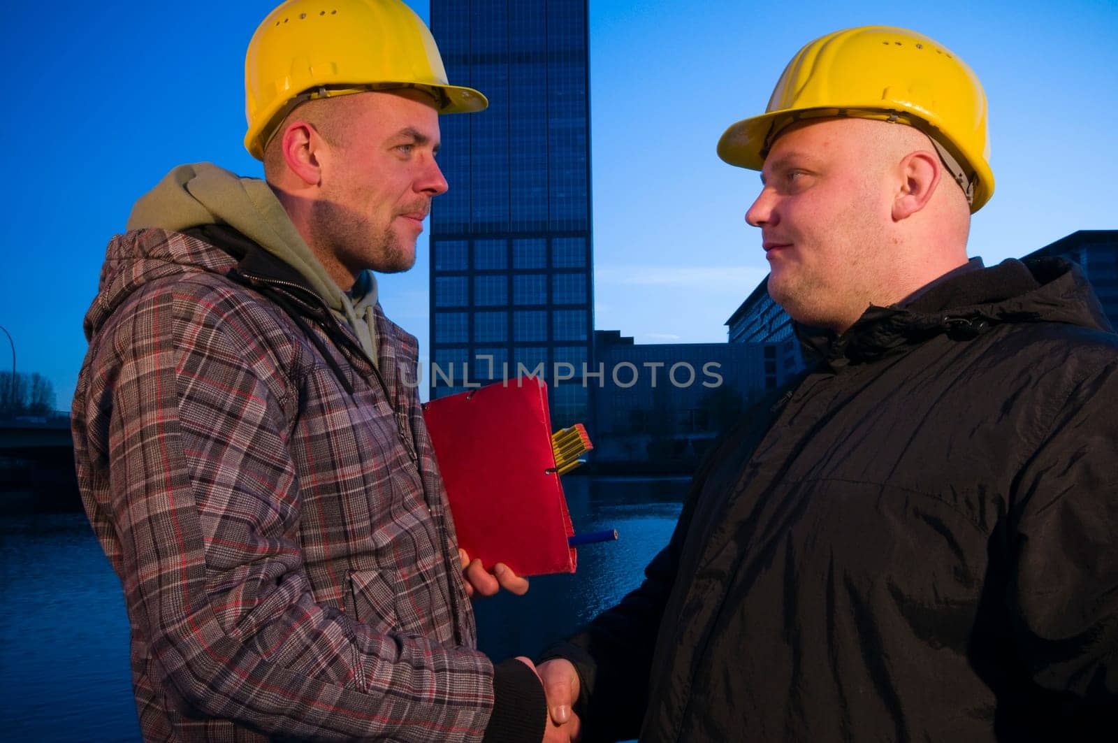 Upper body shot of two men with yellow construction site helmets standing opposite each other and shaking hands and looking into each other's eyes