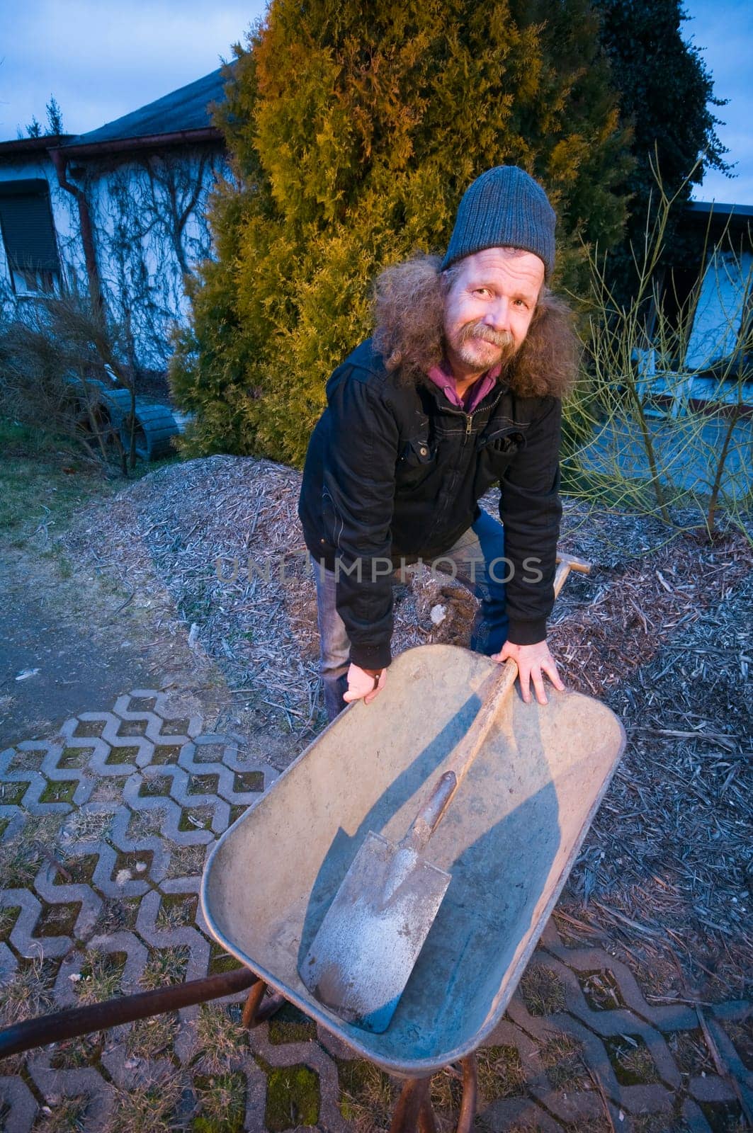 Wide-angle shot of an elderly man bent over with a shovel and a shovel lying in it at dusk and looking into the camera