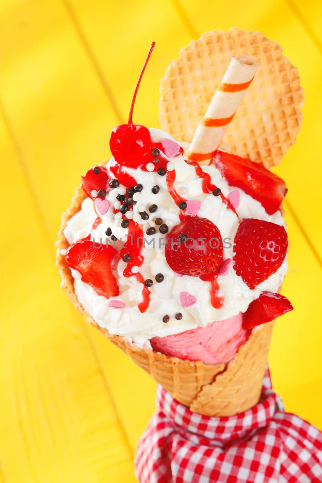 Colourful party ice-cream cone garnished with ripe red strawberries and a spiral wafer biscuit on a cheerful yellow background