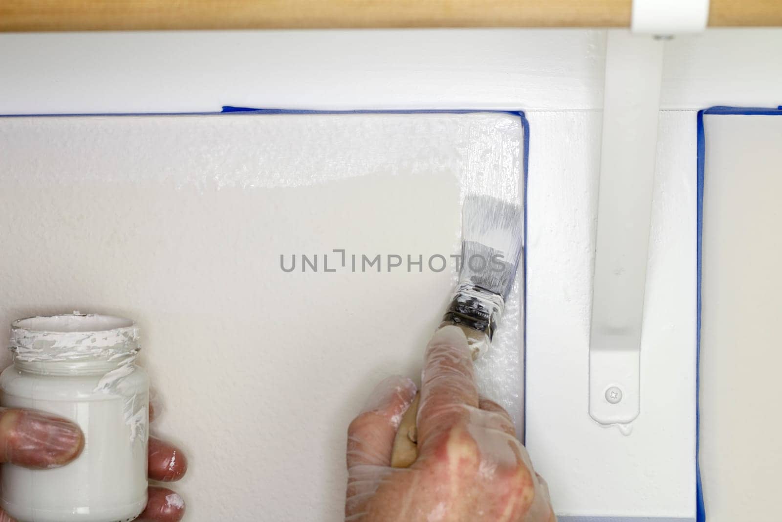 Hand holding a glass jar of white paint in left hand while painting a corner of a wall under a shelf in a closet close up.