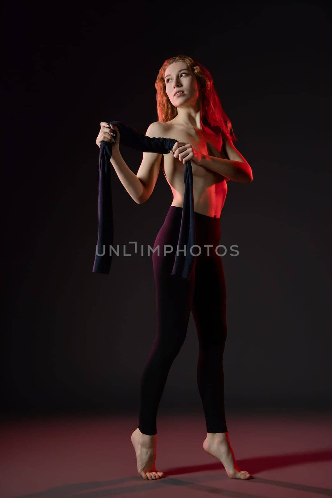 A woman showcases her beauty while dancing topless, holding a dark fabric, illuminated by artistic lighting in a studio setting.