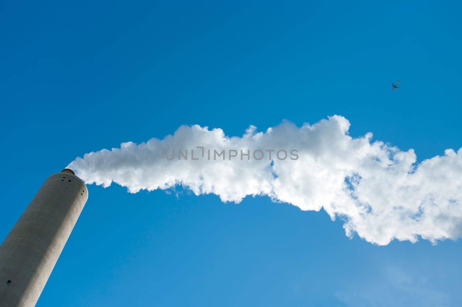 smoking industrial chimney against a blue sky with an airplane passing by