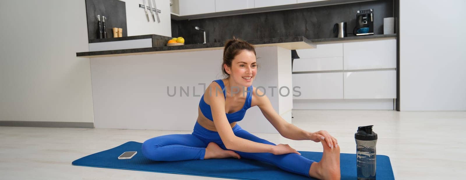 Fitness and workout concept. Young woman stretching her legs, doing exercises at home on yoga mat, doing splits on floor in living room by Benzoix