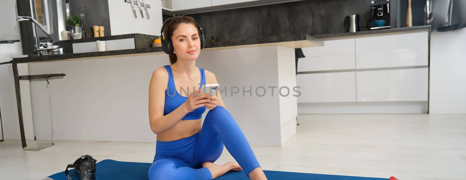 Portrait of young happy woman does workout in headphones, listens to music while doing yoga, sitting on rubber mat in blue sportswear.