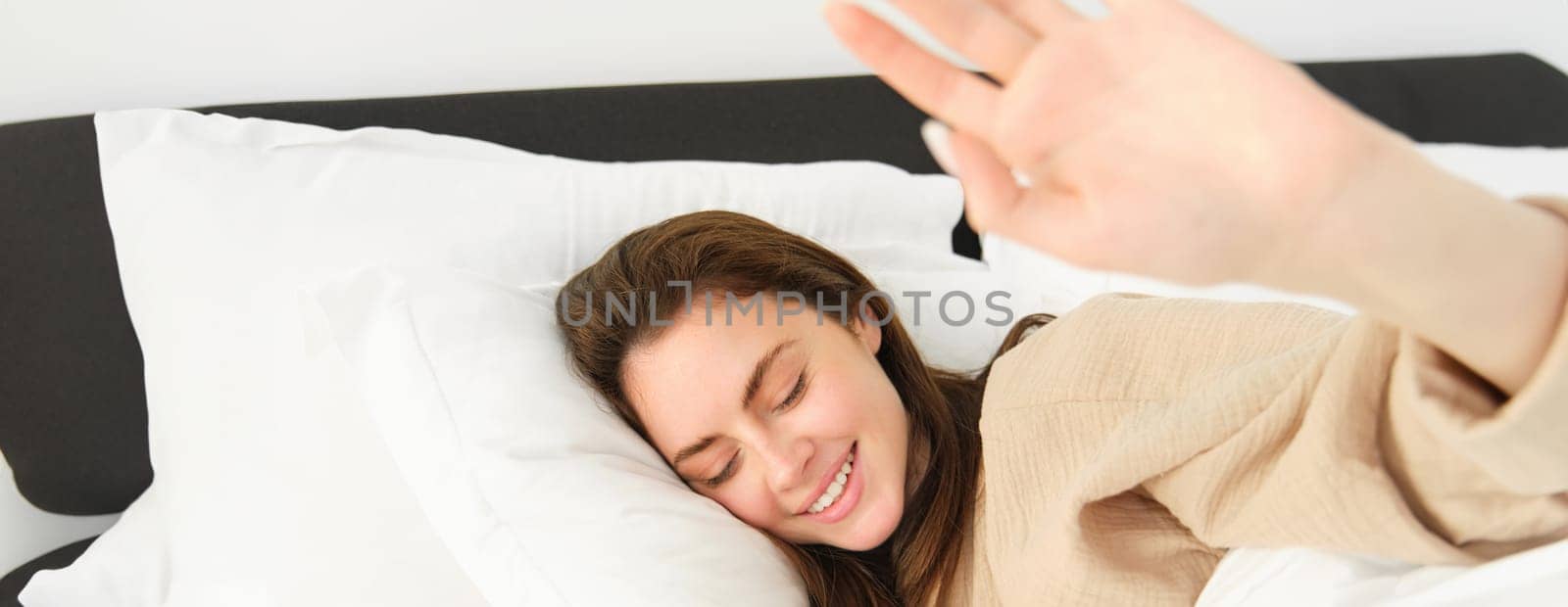 Portrait of gorgeous girlfriend stretching hand, blocking camera, lying in bed and resting on pillow, smiling while waking up from someone by Benzoix