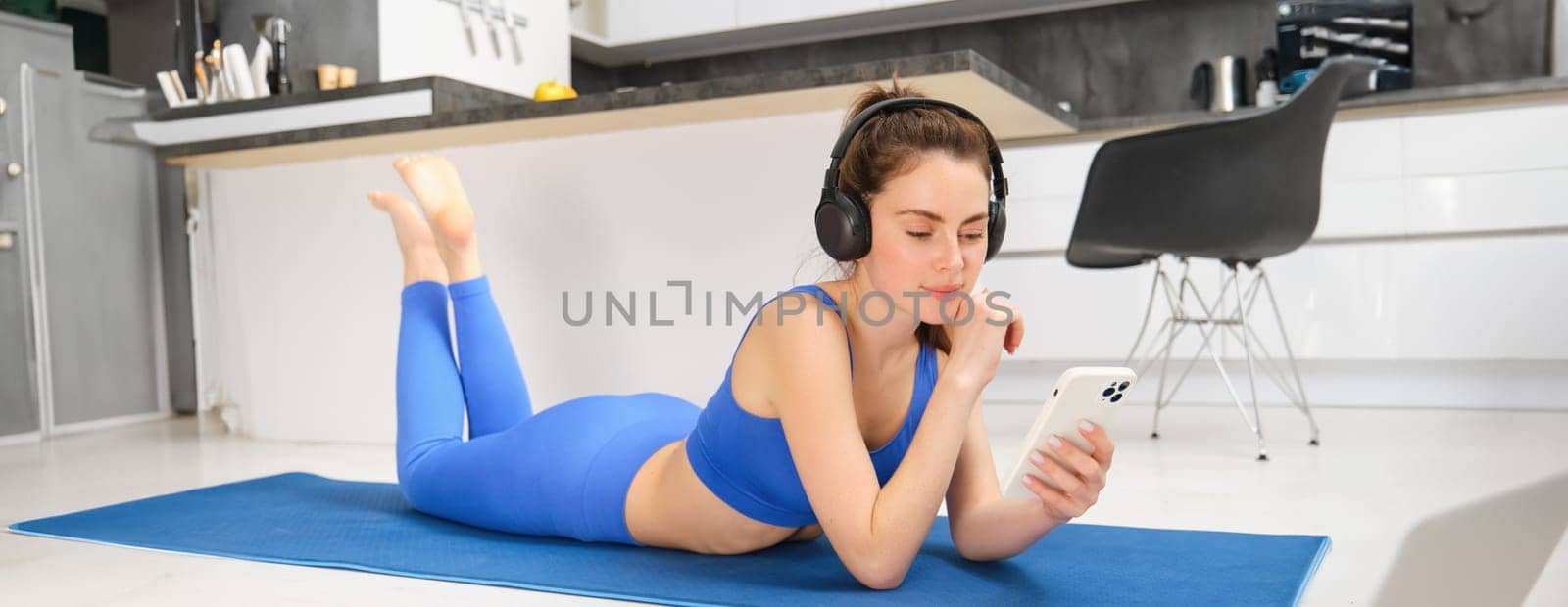 Image of young woman watches sport gym tutorials on smartphone, laying on yoga mat, doing fitness workout at home, wearing blue leggings and sportsbra.