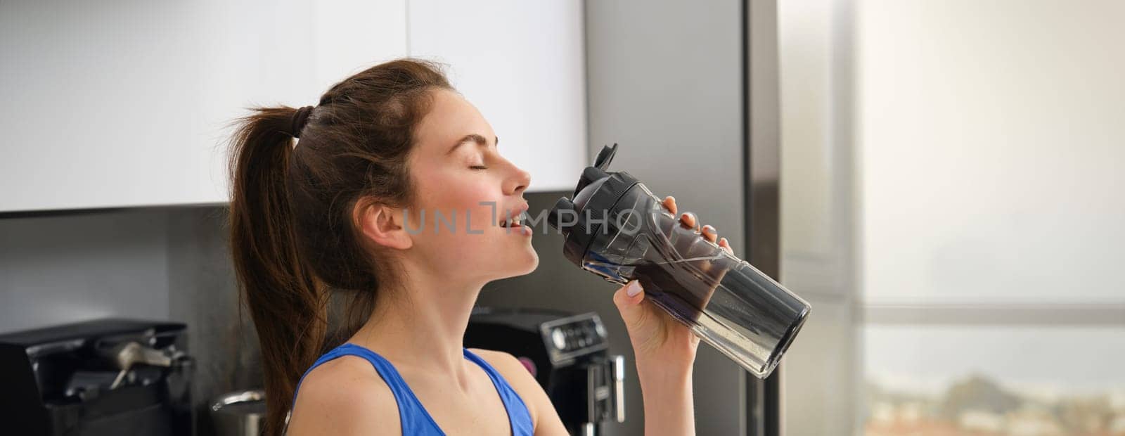 Close up portrait of sportswoman face, fitness girl drinks water from sport bottle and smiling, hydrating after productive aerobics, yoga workout.