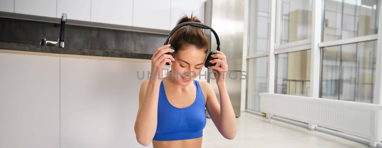 Young woman doing workout training at home, puts on her wireless headphones by Benzoix