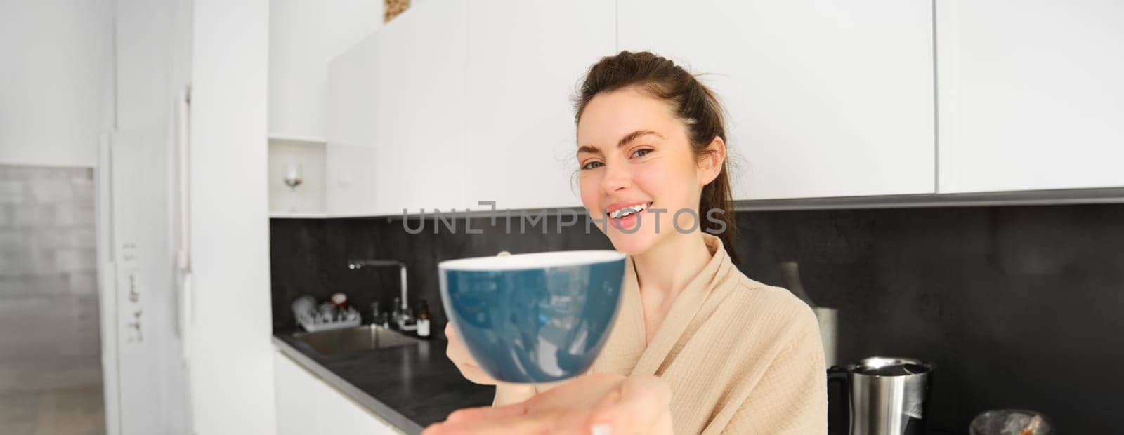 Portrait of attractive smiling woman giving you cup, offering morning coffee and looking happy, standing in the kitchen in bathrobe by Benzoix