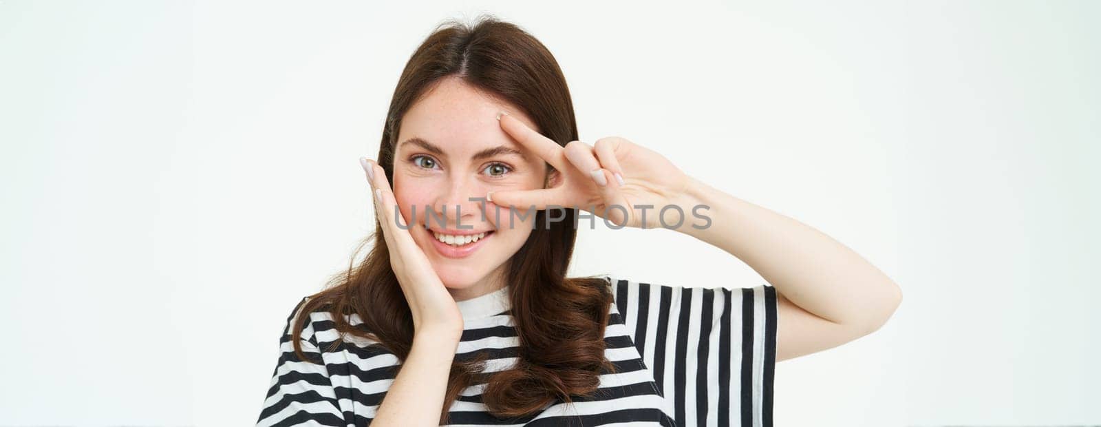 Portrait of beautiful, smiling female model, touches her clear glowing skin, shows peace sign and looks happy at camera, isolate on white background.