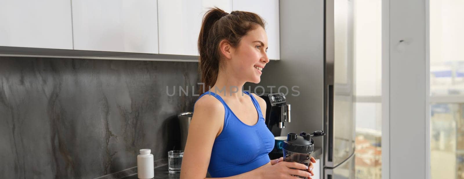 Fitness woman standing with black water bottle in kitchen, wearing workout clothing, drinking after yoga exercises.