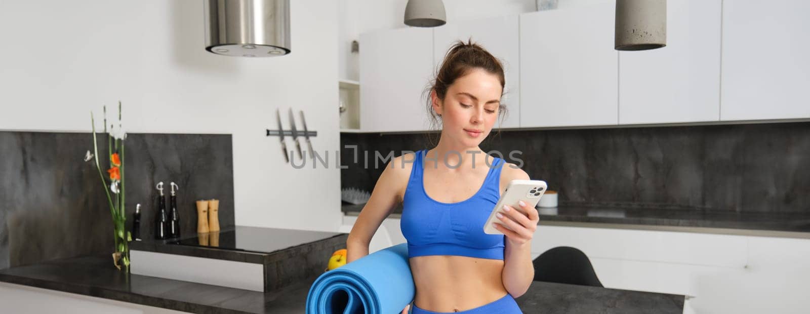 Portrait of attractive, fit young woman in sportswear, standing in living room with rubber yoga mat, holding smartphone, doing fitness exercises with mobile app at home by Benzoix