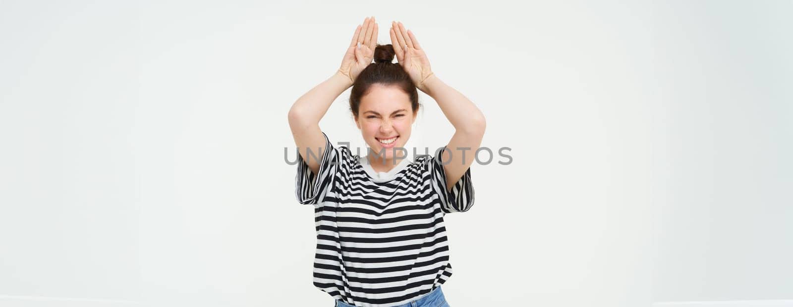 Image of beautiful, happy young woman showing bunny ears on top of her head, looking excited, posing over white background. Lifestyle and people concept