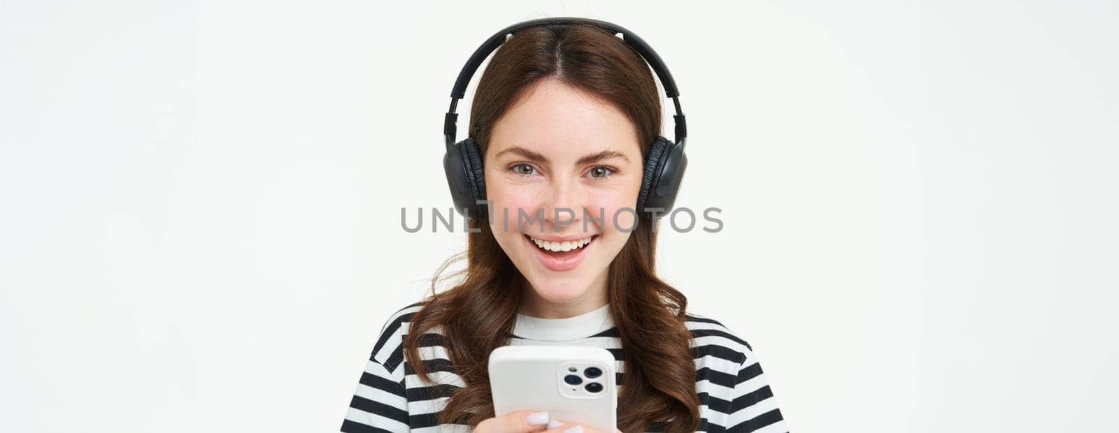 Image of young woman in headphones, using smartphone and laughing, watching video on mobile phone, listens to music on streaming service app, white background.