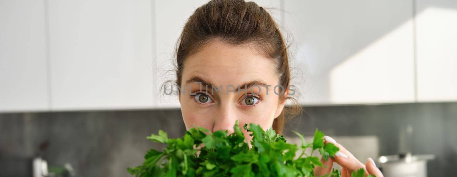 Portrait of attractive girl with bunch of parsley, eating fresh herbs and vegetables, cooking in the kitchen by Benzoix