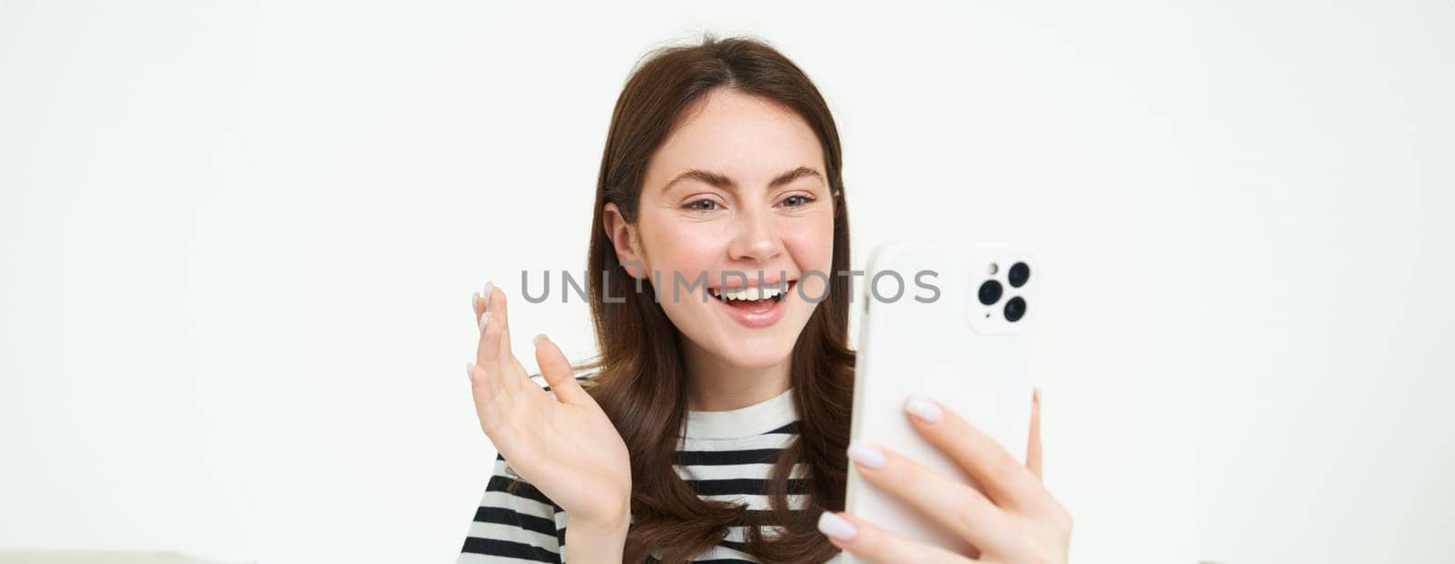 Portrait of young modern girl connects to video chats, talks with friend online using smartphone app, waves at mobile phone camera, white background.