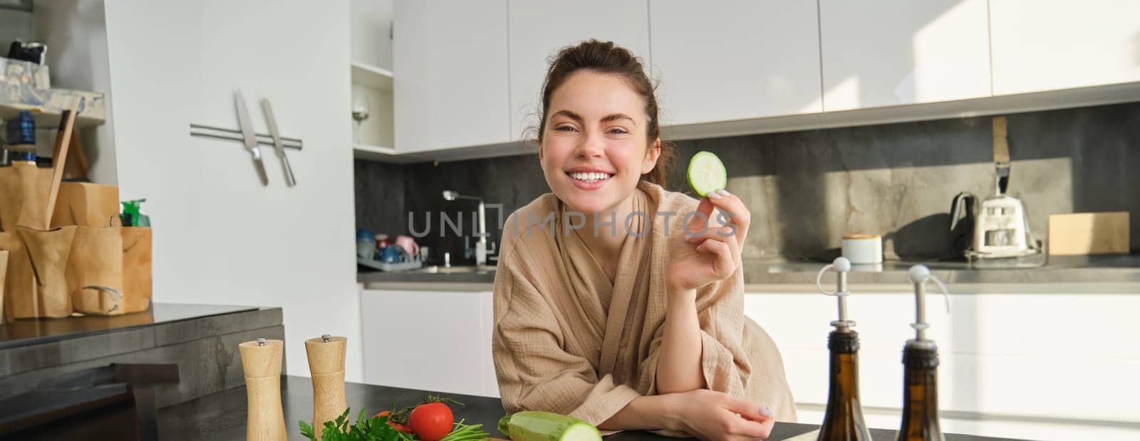 Portrait of cute young woman in bathrobe, cooking meal, standing near chopping board with vegetables, holding zucchini, making salad or vegetarian dinner.