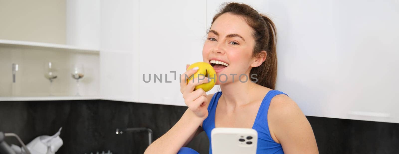 Portrait of smiling brunette girl, wearing fitness clothes, sitting in kitchen and biting an apple, eating fruit snack between workout, holding smartphone, using mobile phone app.