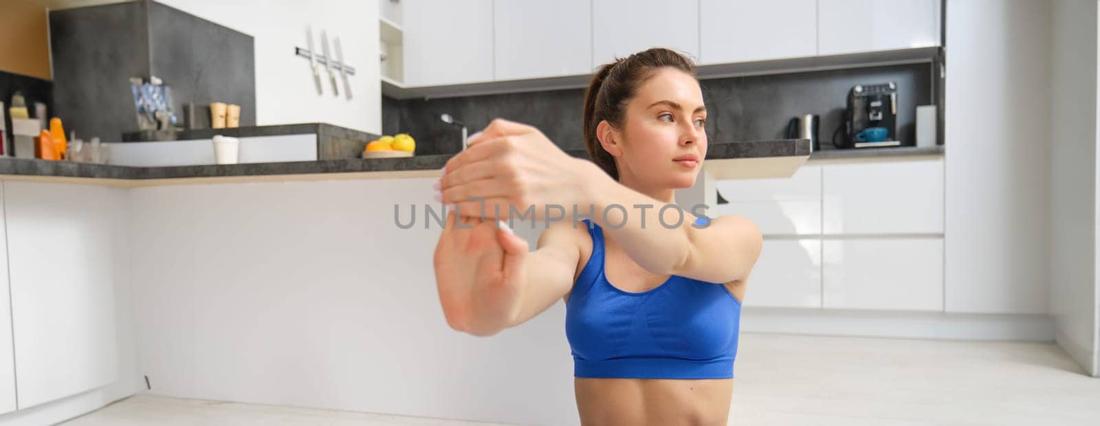 Portrait of beautiful woman does fitness training, sits on floor near kitchen, stretching her hands, workout from home. Copy space