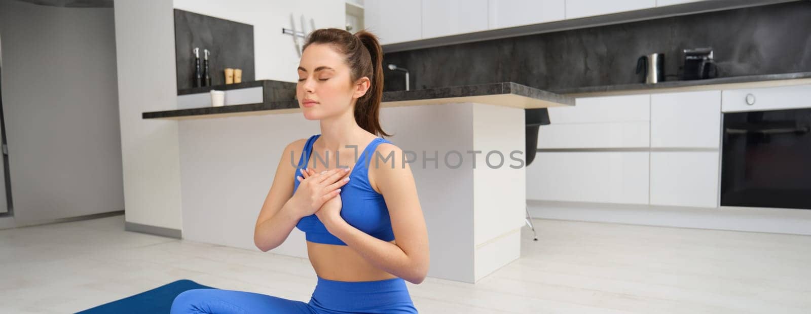 Side show of young woman meditating at home, wearing blue sportswear, sitting in lotus pose on rubber yoga mat, holding hands on chest, controlling breathing.