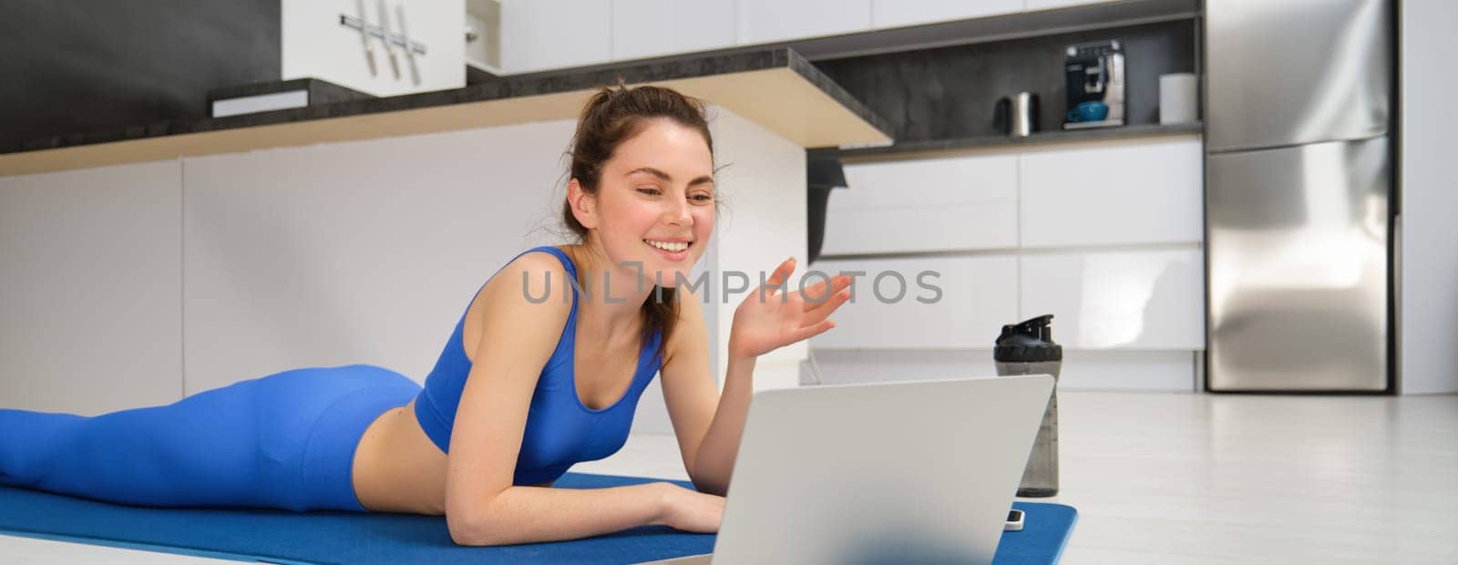 Image of young fitness girl talking online on laptop, video chats, laying on floor rubber mat, doing exercises, discuss workout training, yoga from home.