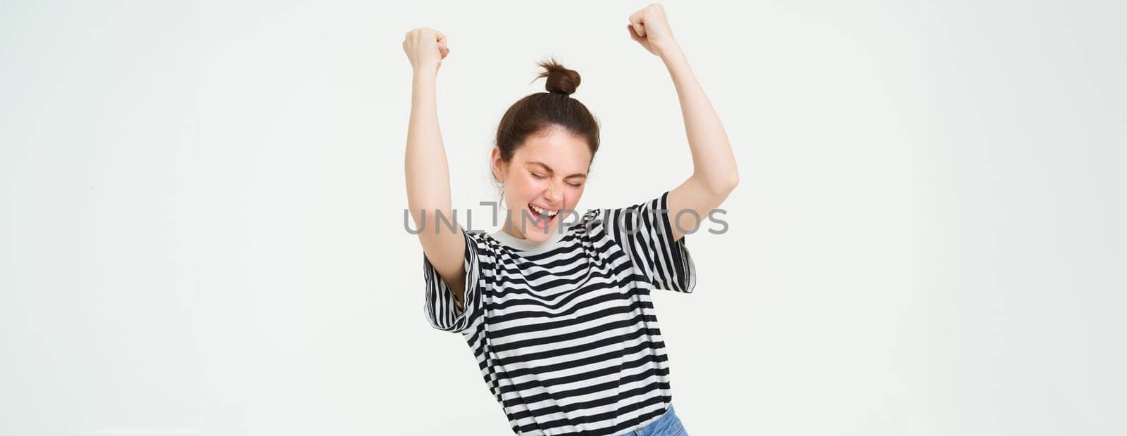 Portrait of happy young sports girl, fan rooting for team, celebrating victory, raising hands up, chanting, triumphing, standing over white background by Benzoix