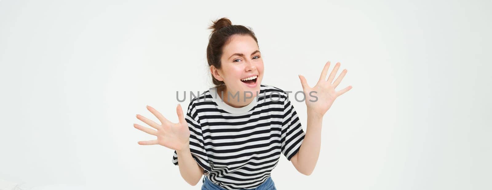 Image of happy, beautiful young woman makes jazz hands, surprise gesture, posing in casual t-shirt and jeans over white background by Benzoix