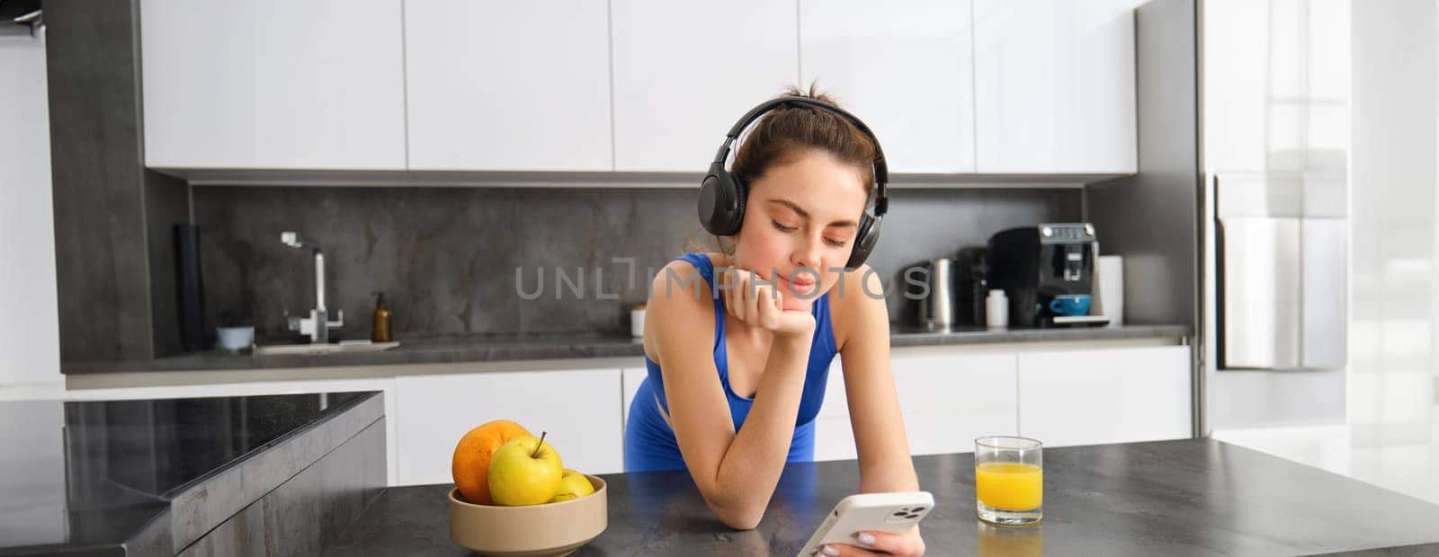 Image of happy, stylish young sports woman, standing in kitchen and drinking orange juice, listening music in headphones, using smartphone app by Benzoix