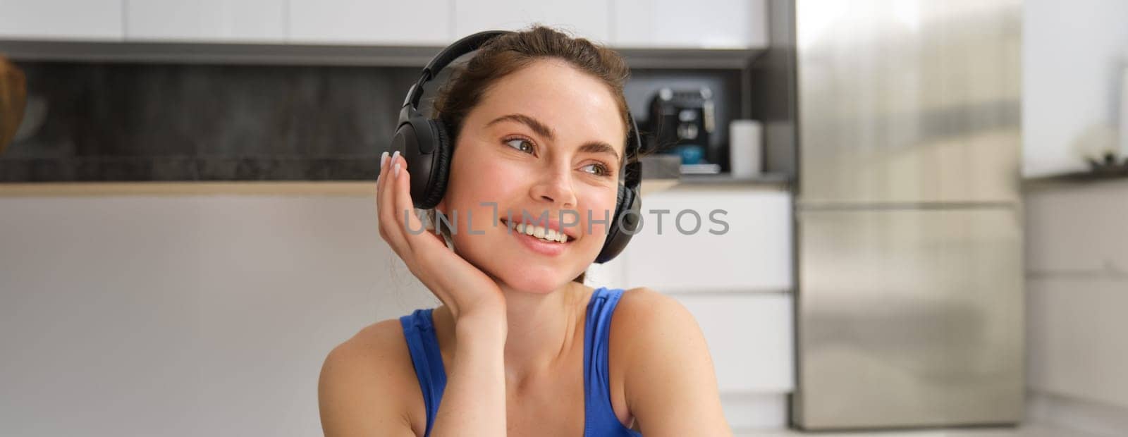 Close up portrait of brunette fitness girl, listens music in wireless headphones, smiles at camera, does workout alone at home, sits on floor and exercises by Benzoix