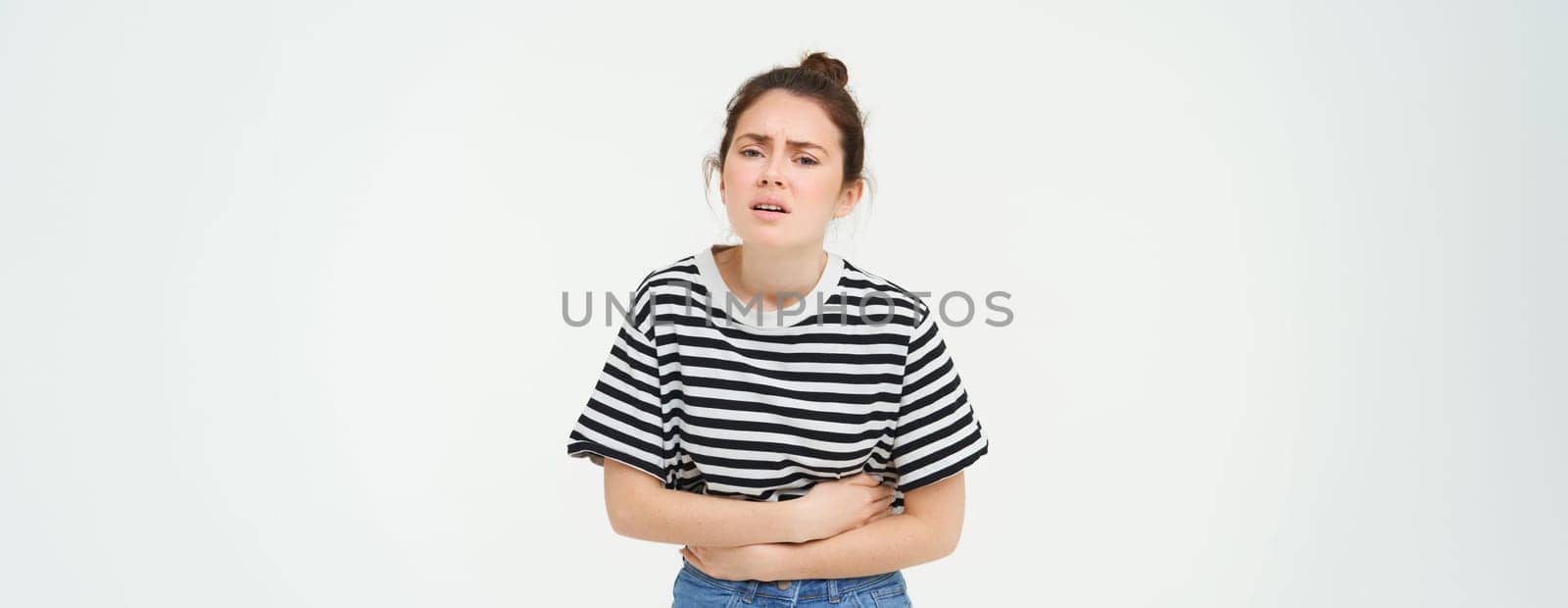 Image of young woman holding hands on her belly, feeling discomfort, menstrual period cramps, needs painkillers, stands over white background.