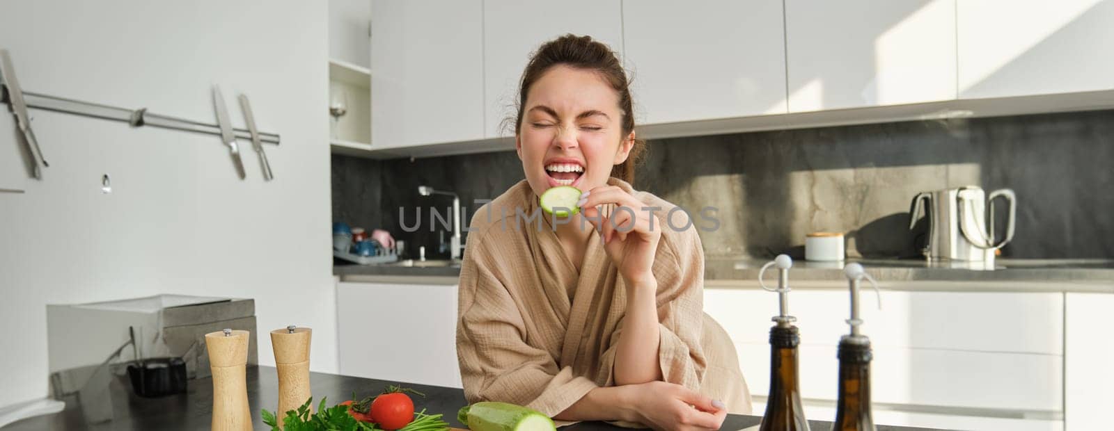 Portrait of happy girl eating vegetables while making meal, cooking healthy vegetarian food in kitchen, holding zucchini, posing in bathrobe by Benzoix