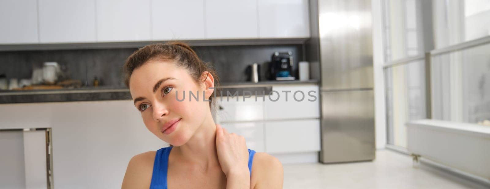 Close up portrait of young woman does fitness at home, touches her neck, rubs shoulders after workout training session. Wellbeing and sport concept