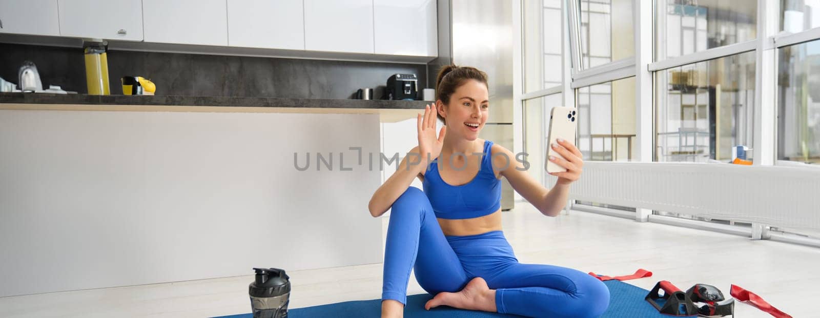 Image of beautiful, atheltic girl sits on yoga mat at home with smartphone, takes selfie and workout, does fitness exercises by Benzoix