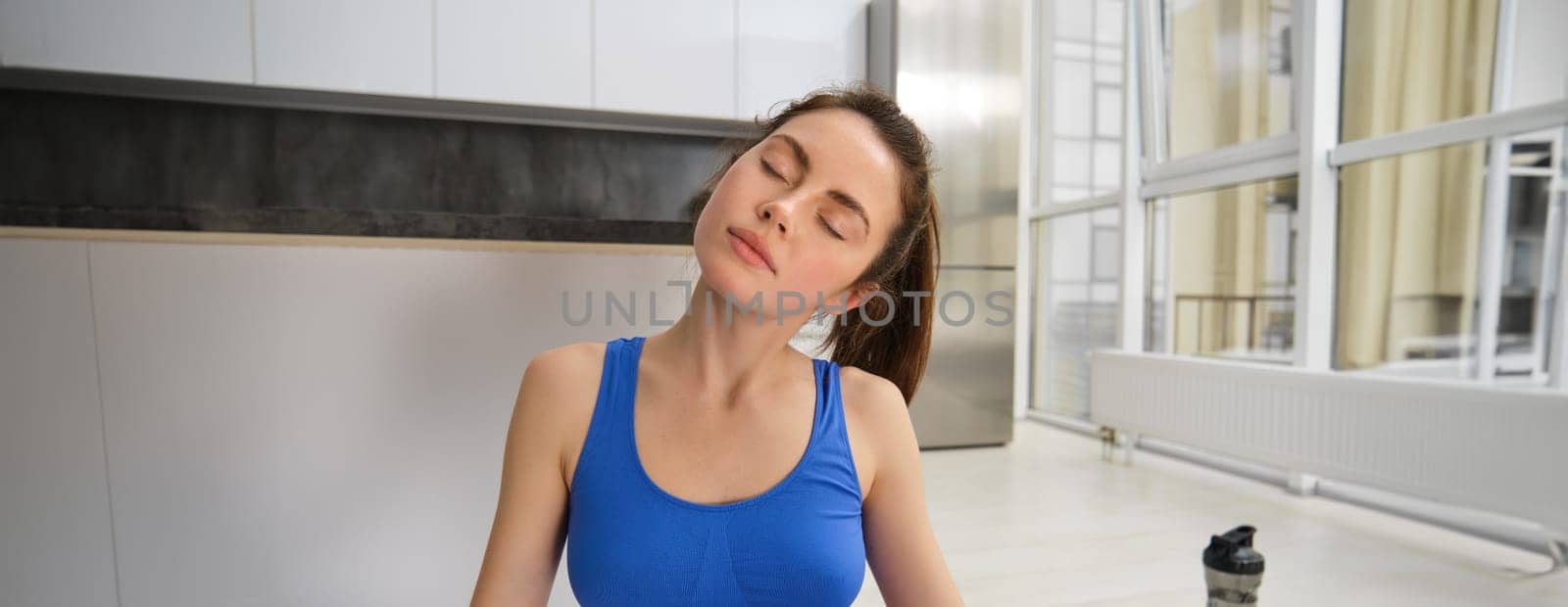 Portrait of young sportswoman, stretching her neck, warm-up before yoga exercises, doing fitness workout on rubber mat, wearing blue sportsbra and leggings by Benzoix