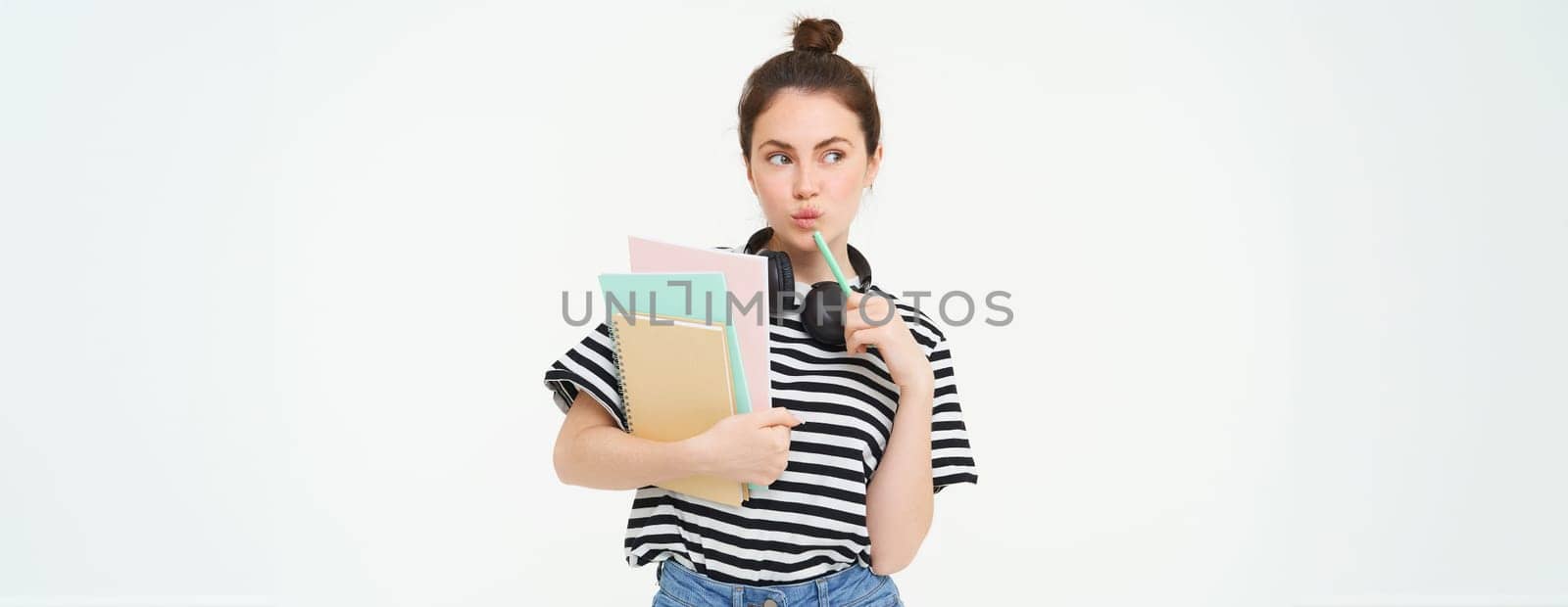 Student and education concept. Young woman with books, notes and pen standing over white background, college girl with headphones over neck posing in studio.