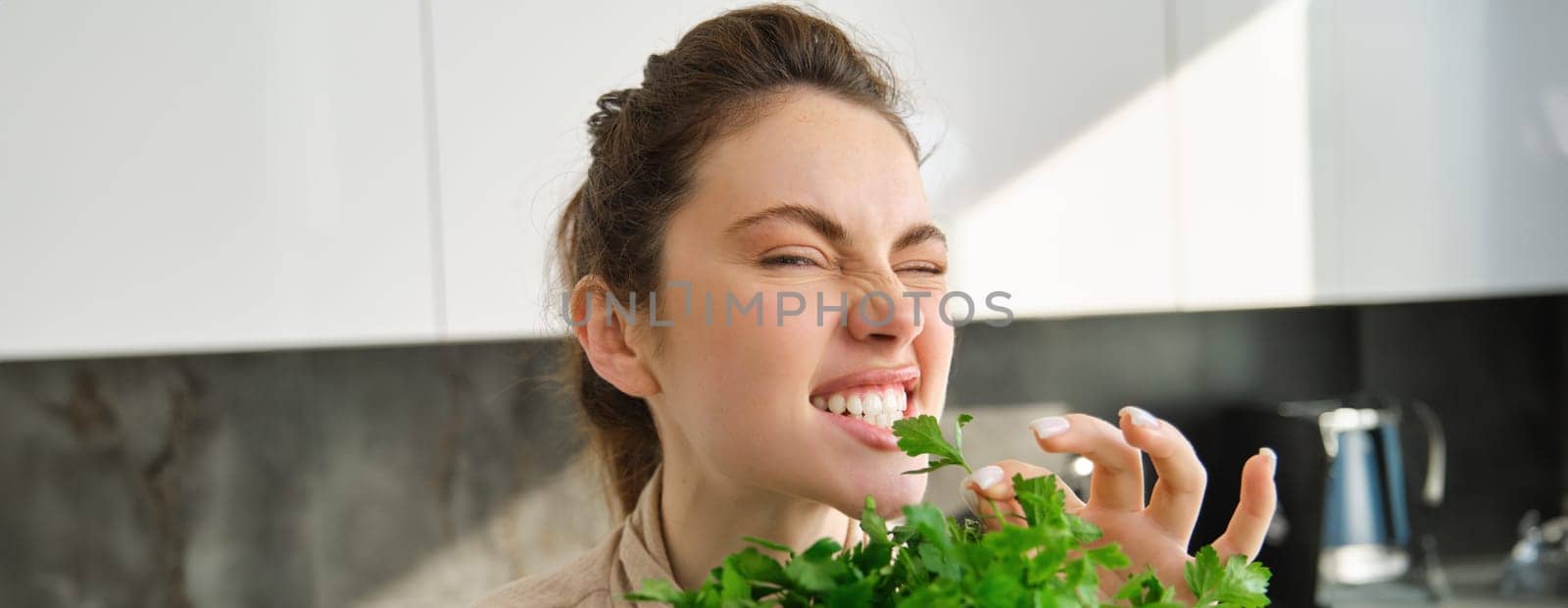 Funny and cute brunette woman bites delicious, fresh green parsley, eating herbs, cooking salad in kitchen by Benzoix